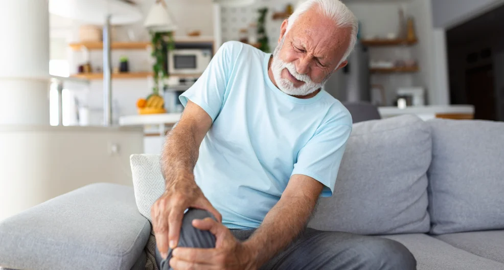 An older man is sitting on the couch at home, and is holding his leg. It looks like he has pain in the knee joint.